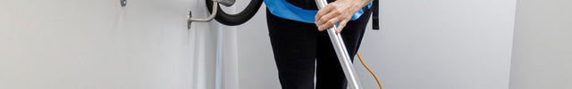 A woman in a blue uniform vacuums a carpeted staircase with a backpack-style vacuum cleaner in a bright white stairwell.