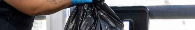 A woman in a blue shirt and dark vest wearing blue gloves empties a black plastic bag into a recycling bin next to a window.