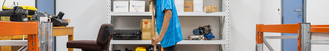 A man in a blue shirt mops the floor in a warehouse or storage room with orange metal racks, shelving, and a yellow caution sign that reads 'CAUTION WET FLOOR / CLEANING IN PROGRESS'.