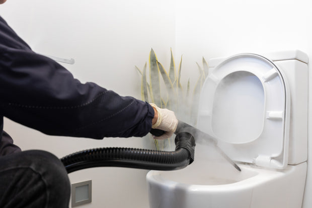 Hands holding steam cleaning machine, steaming the ubnderside of toilet seat to sanitise and remove grime