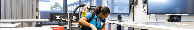 A person in a blue cleaning shirt stands in a modern classroom or training room with multiple large computer monitors, white desks, and blue-gray carpeting. Vacuuming