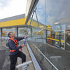 A worker in a high-visibility orange vest cleans a large glass storefront window with a long-handled squeegee, with a yellow and gray building in the background.